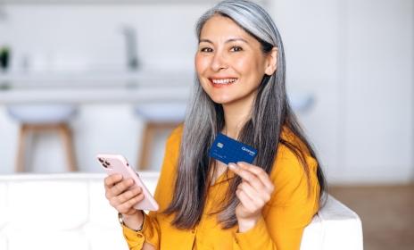 A woman sitting in her living room holding a Quorum Debit Mastercard and her smartphone, smiling at the camera.
