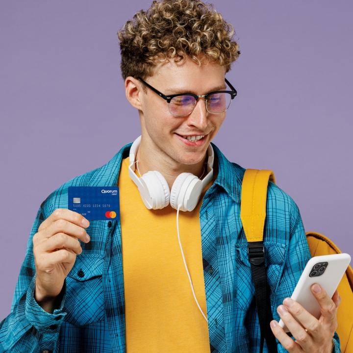 A college student holding a Quorum debit card, while looking at Quorum's mobile banking app.